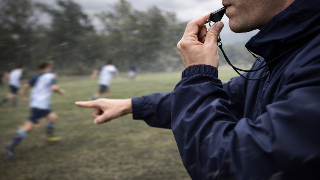 強風の屋外グラウンドでコーチが笛を吹いて合図を出すイメージ写真